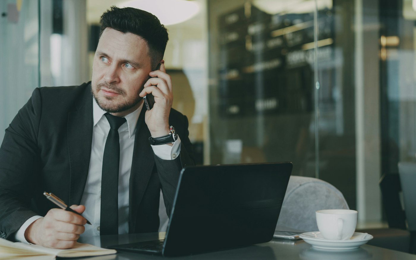 A sales rep on a call at a desk, taking notes beside a laptop